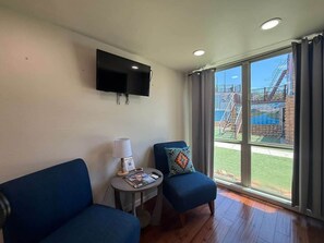 A room with a flat-screen TV mounted on the wall, a small table with a lamp and a magazine, two blue armchairs, and a large window with a view of an outdoor staircase and greenery.
