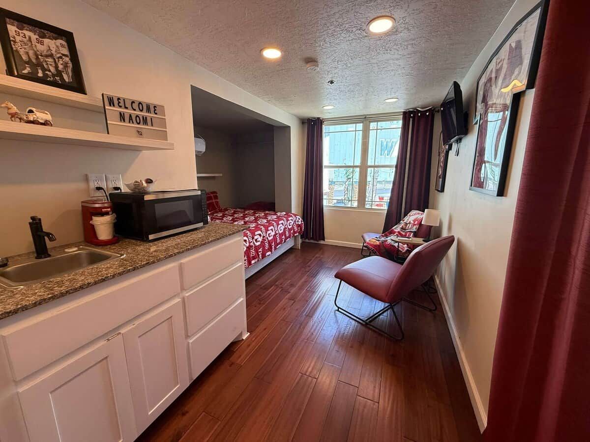 Eine kompakte Küche mit Mikrowelle, Spüle und Schränken. Im Hintergrund ist ein Bett mit einer rot gemusterten Decke zu sehen.
