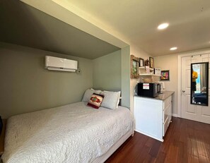 A bedroom with a bed, a small kitchenette, and a door leading to another room.