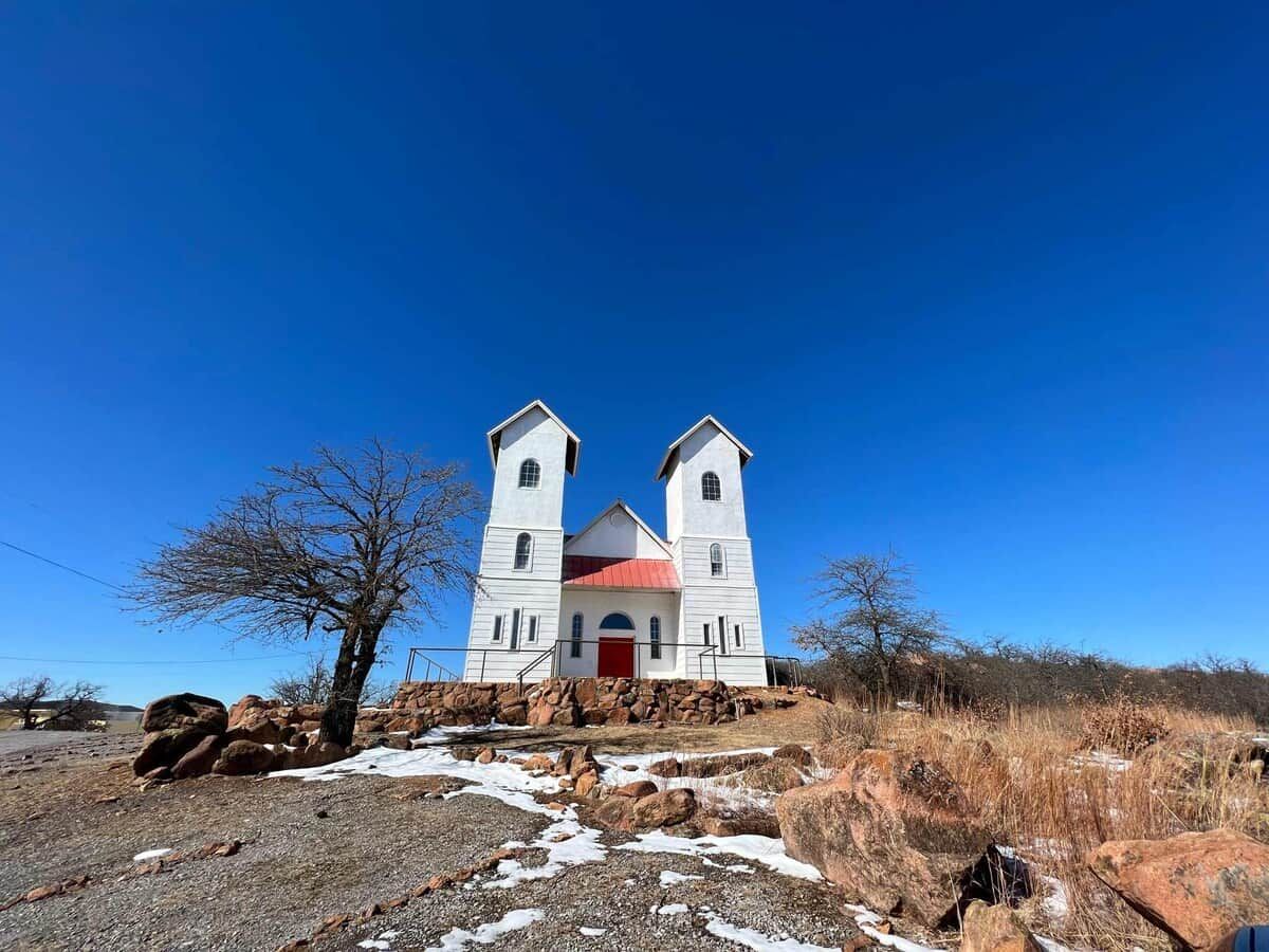 A white church with a red door and two towers, surrounded by dry grass and rocks.