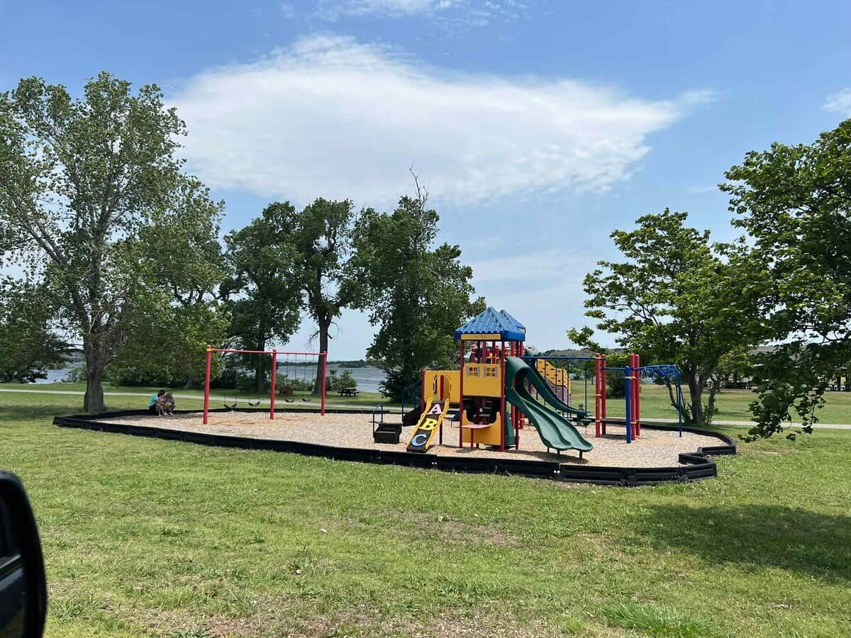A playground with a slide and climbing structures, surrounded by grass and trees.
