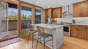 Una cocina moderna con armarios de madera, una isla grande con taburetes de bar, una estufa de acero inoxidable y vistas a una montaña nevada a través de amplios ventanales.