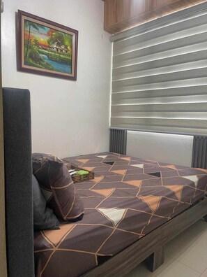 Ein Schlafzimmer mit Bett, Kissen, Nachttisch mit Buch, gerahmtem Bild an der Wand und Fenster mit Jalousien.
