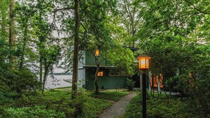 A pathway leading to a green building with a visible door and windows, surrounded by lush greenery and a body of water in the background. - Scenic, Spacious Riverside Retreat | Ropeyarn (Deltaville)