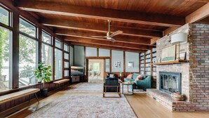 A spacious living room with a fireplace, large windows, and a ceiling fan. - Scenic, Spacious Riverside Retreat | Ropeyarn (Deltaville)