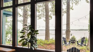 A room with large windows overlooking a lake, featuring wooden benches and a view of outdoor seating with chairs and a fire pit. - Scenic, Spacious Riverside Retreat | Ropeyarn (Deltaville)