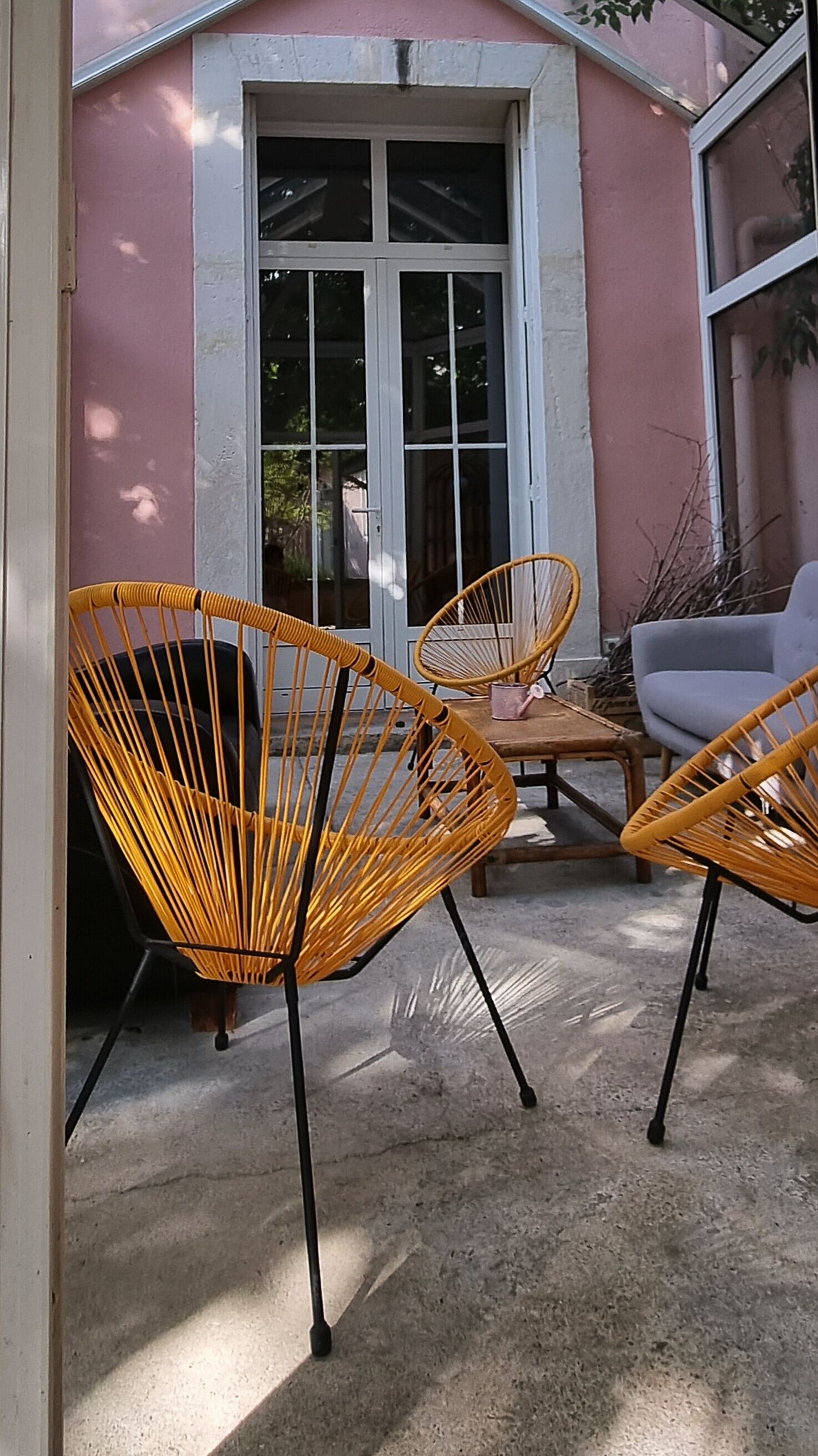 A patio area with wicker chairs and a table, a pink building with a large window, and a concrete floor.