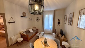 Un salon chaleureux avec un canapé en bois, une table à manger ornée d’un petit sapin de Noël, un téléviseur et une grande fenêtre avec des rideaux.