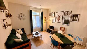 A cozy living room with a sofa, a dining table, and a TV. There are framed pictures on the wall and a clock. - Villa Bleue 3 Le Mont Dore 5pers (Mont-Dore)