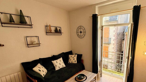 A cozy living room with a dark green sofa, a wooden coffee table, and a small wooden stool. There are wall-mounted shelves with decorative items and a window with curtains. - Villa Bleue 3 Le Mont Dore 5pers (Mont-Dore)