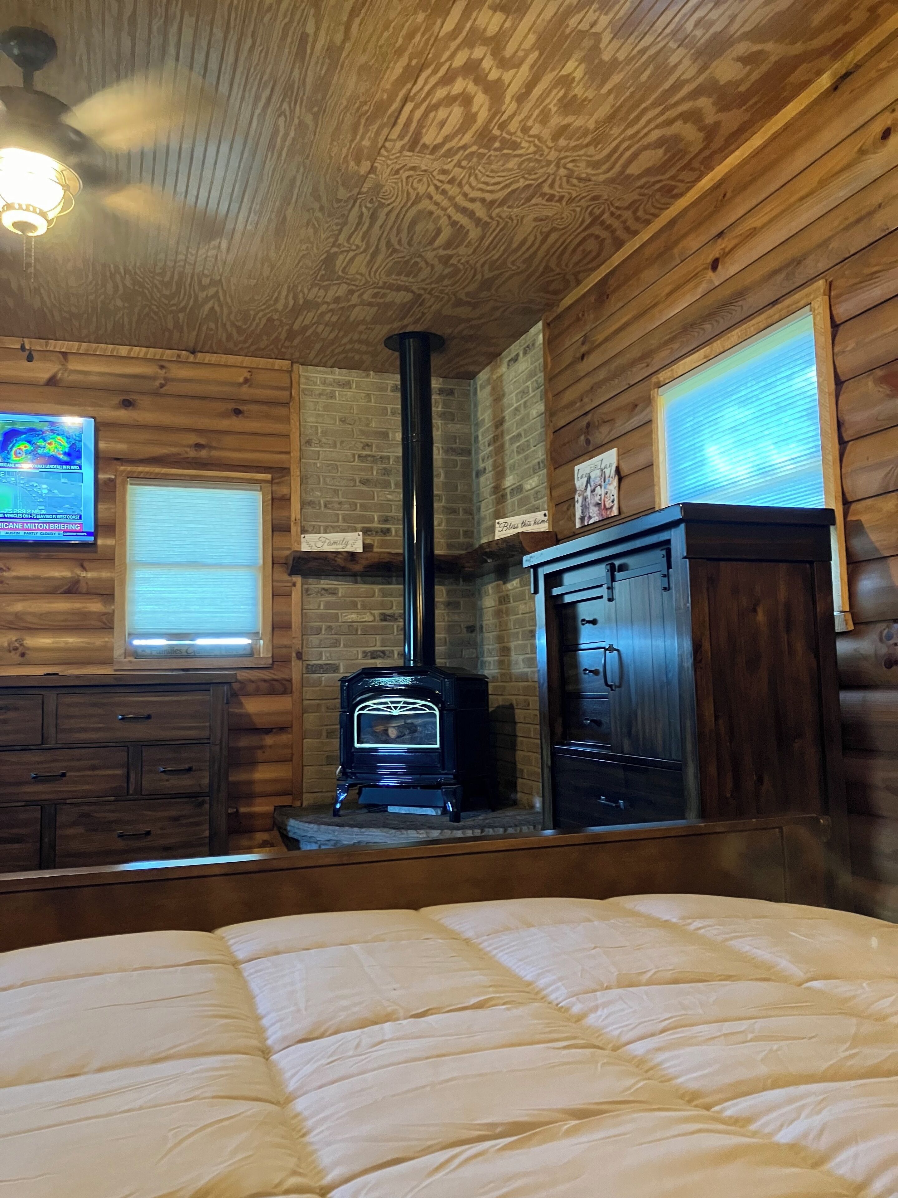 A log cabin room with a wooden interior, a black wood stove, a wooden dresser, and a bed with a white comforter.