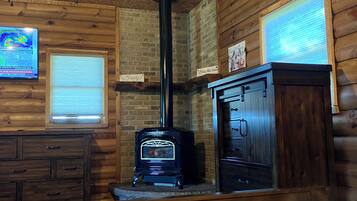 A log cabin room with a wooden interior, a black wood stove, a wooden dresser, and a bed with a white comforter.