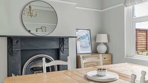 Un comedor con una mesa de madera, sillas blancas, chimenea, espejo y ventana con persianas.