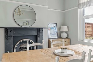 Un comedor con una mesa de madera, sillas blancas, chimenea, espejo y ventana con persianas.