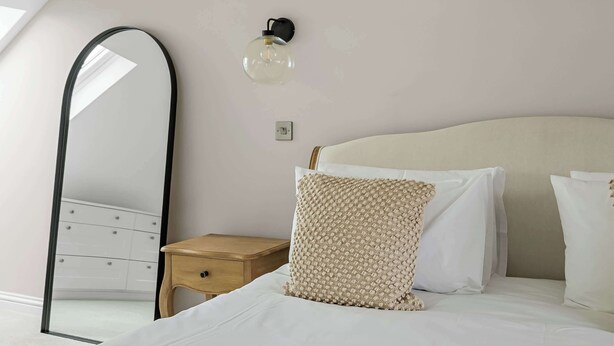 Un dormitorio con una cama, una mesita de noche de madera, un espejo y una lámpara fijada a la pared.