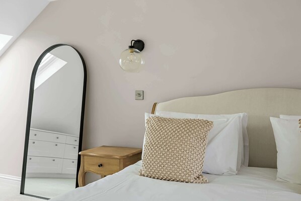 A bedroom with a bed, a wooden nightstand, a mirror, and a wall-mounted light fixture.