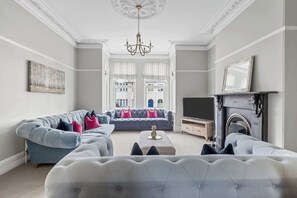 A living room with a grey sofa, a tufted ottoman, a fireplace, and a chandelier.