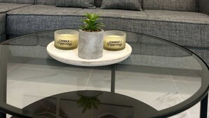 Ein Wohnzimmer mit einem grauen Sofa, einem Glastisch mit drei Kerzen mit dem Aufdruck 'CANDLE of Happiness' und einem kleinen Blumentopf.