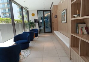 A modern lounge area with blue chairs, a wooden bookshelf, and a view of the outside balcony.