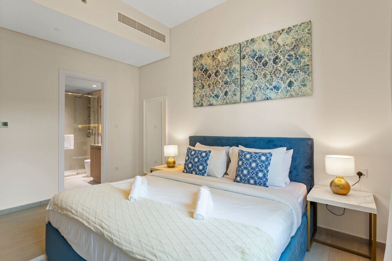 Una habitació d'hotel amb un llit gran, un panell del capçal blau, dues taules de nit amb làmpades i una obra d'art a la paret sobre el llit.