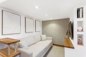 A modern living room with a white sofa, a wooden coffee table, and a flat-screen TV mounted on the wall. There are abstract paintings on the wall and a small shelf with decorative items.