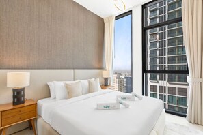 A hotel room with a large bed, two bedside tables with lamps, a view of a cityscape through large windows, and a curtain.