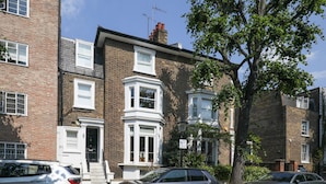 A row of terraced houses with a central bay window, a tree in the foreground, and parked cars along the street. - Charming 1BR Flat in Quiet S Kensington (London)