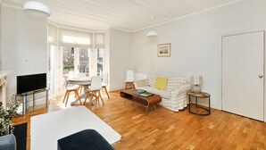 A spacious living area with a wooden floor, a sofa, a coffee table, a TV on a stand, and a dining table with chairs. - Charming 1BR Flat in Quiet S Kensington (London)