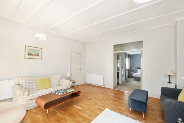 A living room with a sofa, a coffee table, and a blue ottoman. There is a door leading to a bedroom. - Charming 1BR Flat in Quiet S Kensington (London)