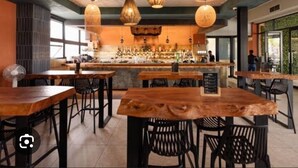 Ein Restaurantinneres mit Holztischen und -stühlen, hängenden Pendelleuchten und einem Barbereich im Hintergrund.