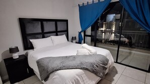 Ein ordentlich bezogenes Bett mit weißer Bettwäsche, einem dunklen Kopfteil und einer grauen Tagesdecke. Ein Nachttisch mit Lampe und einem kleinen dekorativen Gegenstand ist vorhanden. Ein großes Fenster mit blauen Vorhängen zeigt eine nächtliche Stadtansicht.