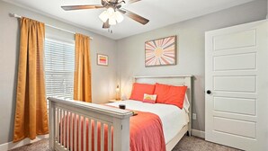 A bedroom with a white crib, a bed with orange bedding, a ceiling fan, and a door.