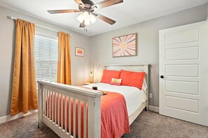 A bedroom with a white crib, a bed with orange bedding, a ceiling fan, and a door.