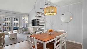 A spacious dining area with a wooden table and white chairs, a large chandelier, and a view of a balcony with outdoor furniture.