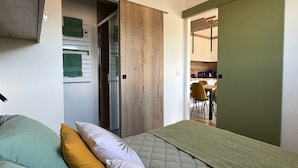 A bedroom with a bed, pillows, a green blanket, a wooden door, and a view into a kitchen area. - Stunning renovated T2, air conditioning, parking, balcony - City and mountain views (Aix-les-Bains)
