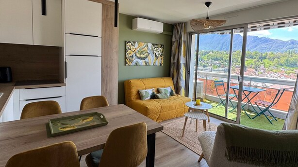 A modern living room with a sofa, dining table, chairs, and a balcony with a view of mountains. - Stunning renovated T2, air conditioning, parking, balcony - City and mountain views (Aix-les-Bains)