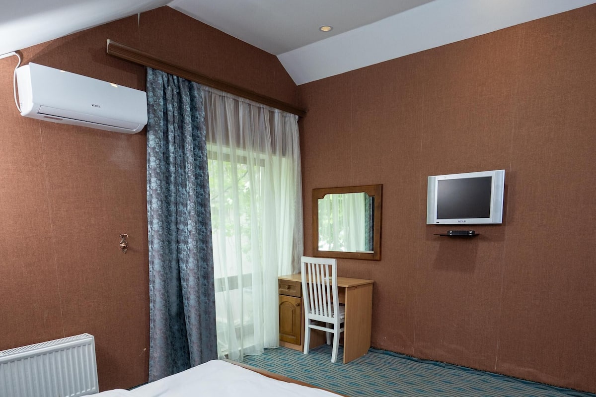 A hotel room with a bed, a desk, a chair, an air conditioner, a television, and a window with curtains.