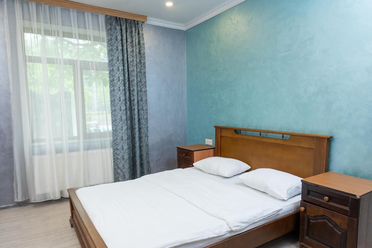 A neatly made bed in a room with a blue wall, a wooden nightstand, and a window with sheer curtains.