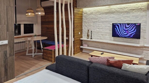 A modern living room with a grey sofa, a wooden dining table, and a TV mounted on a white brick wall. - Cozy apartment in the city center with AC & Wi-Fi (Tashkent)