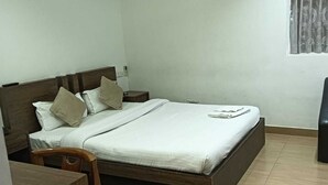A neatly made bed with pillows, a bedside table, a chair, and a window with curtains. - Hotel Vrindavan Palace (Giridih)