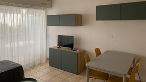 Une pièce moderne et bien éclairée, avec une table à manger, des chaises jaunes, un téléviseur posé sur un meuble en bois et une fenêtre ornée de rideaux légers.