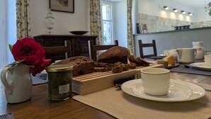 Une table à manger avec un pichet blanc, un pot, une miche de pain, une assiette avec une tasse et un vase contenant une fleur rouge.