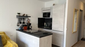 Une cuisine compacte équipée d’un four à micro-ondes, d’une cuisinière et d’un évier. On y trouve un petit îlot central avec une machine à café et des ustensiles. Un canapé jaune est visible au premier plan.