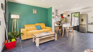 Un appartement moderne avec un canapé jaune, une table basse en bois, une plante rouge et un coin cuisine équipé d’un réfrigérateur et de tabourets de bar.