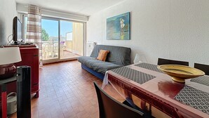 A living room with a sofa, a dining table, and a TV. There is a balcony with a view of the outside. - Bright studio 4 pers, near beach with Wi-Fi (Mauguio)