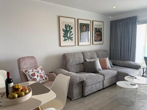 Uma sala de estar moderna com um sofá cinza, uma mesa redonda pequena e uma cadeira. Há impressões botânicas e artísticas emolduradas na parede.