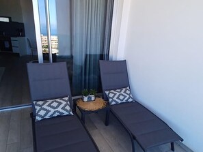 A balcony with two lounge chairs, a small table with a plant, and a view of a cityscape through large windows. - New luxury complex near the sea (Playa Paraiso)