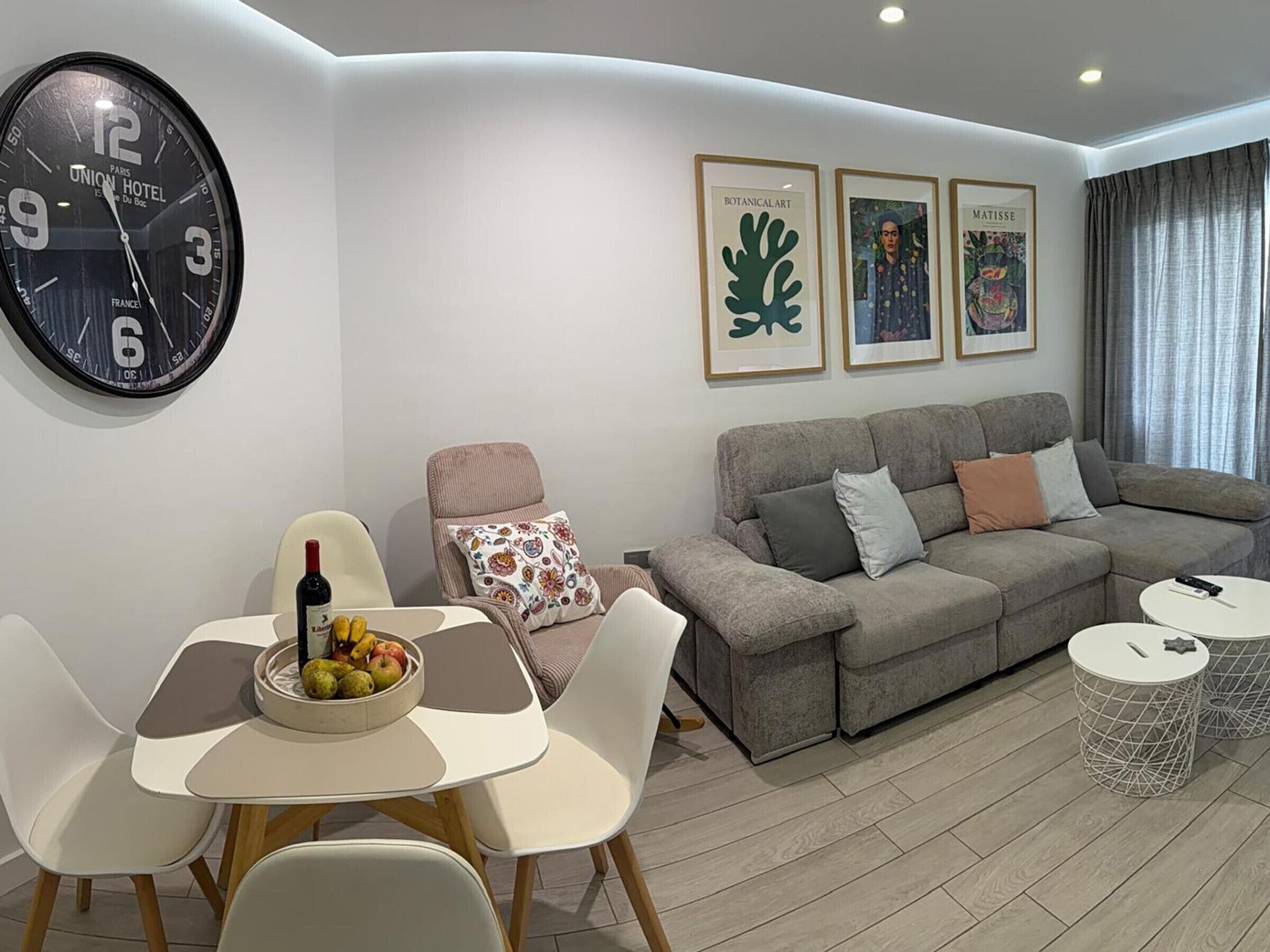 Uma sala de estar moderna com sofá, cadeiras, uma mesa de jantar com frutas, um relógio e obras de arte emolduradas.