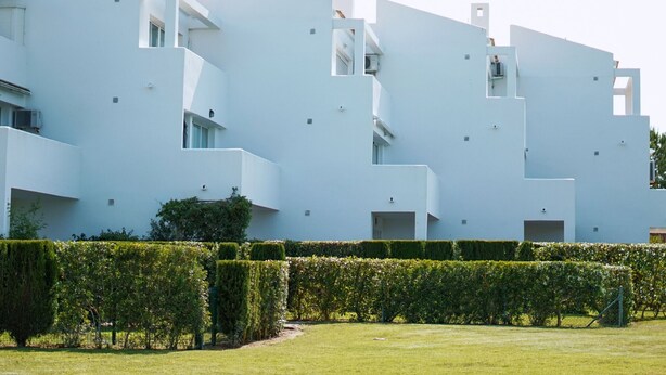 A modern building with a flat facade, multiple balconies, and a well-maintained garden with hedges.