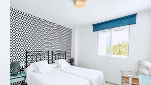 A bedroom with two beds, a desk, a chair, a window with a view, and patterned wallpaper.